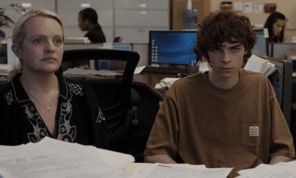 Two office workers sit at a desk with scattered papers, looking ahead in a busy open-plan office.