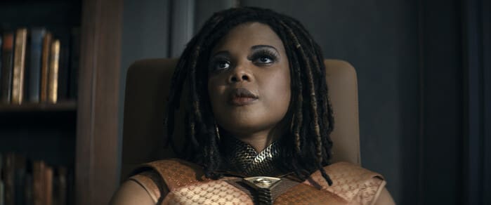 Woman with dreadlocks in ornate orange armor, seated in a high-back chair, looking upward in a dim room.