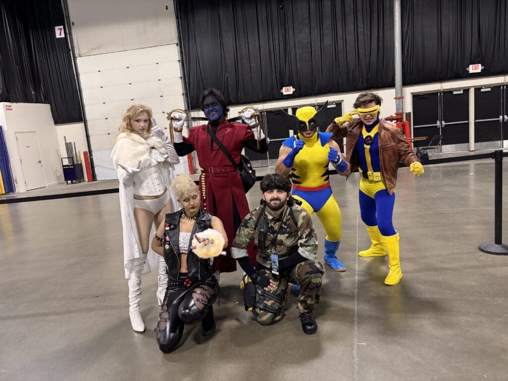 Cosplayers in colorful costumes pose together in a large indoor convention hall, smiling for the camera.