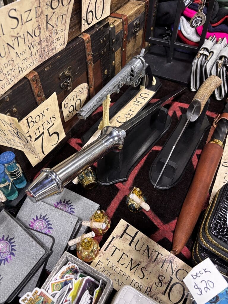 Display of fantasy weapons at a market: ornate silver sword, scabbards, and price signs among wooden chests.
