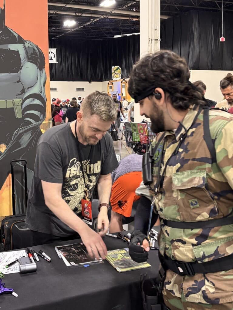 Two men at a convention signing posters at a table; one in camouflage gear with head headset, the other leaning over to sign/draw. In the background, people and vendor booths.