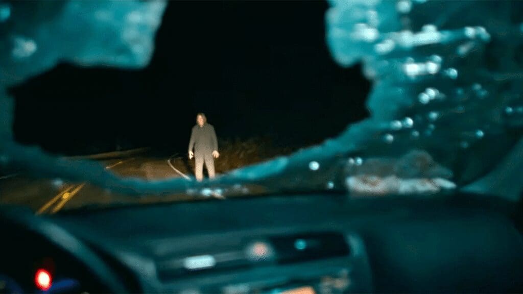 Person in dark clothing stands on a desolate, nighttime road as seen from inside a car with a blue-tinted, cracked windshield framing the view.