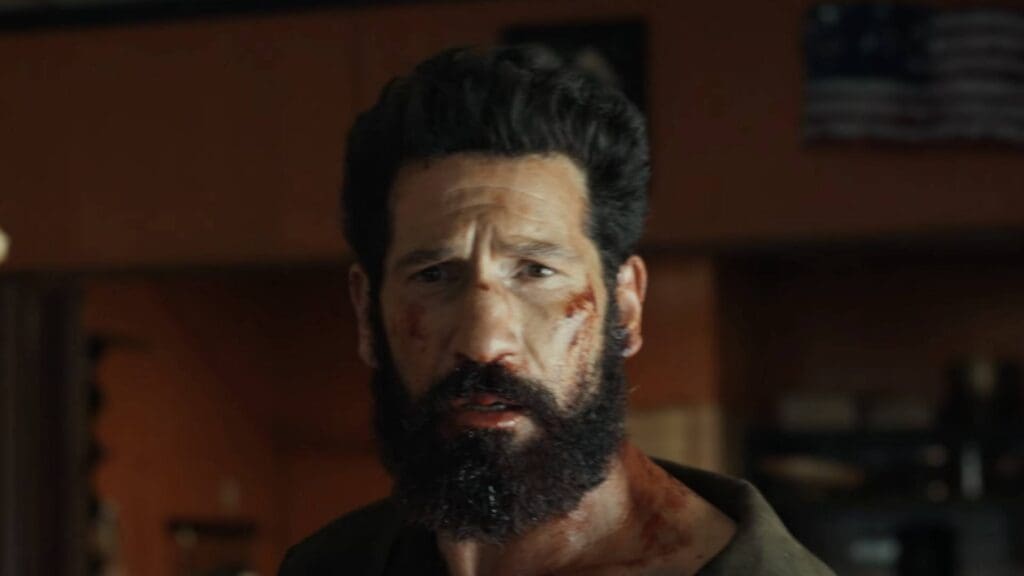 Bearded man with blood on his face looking distressed in a dimly lit room.