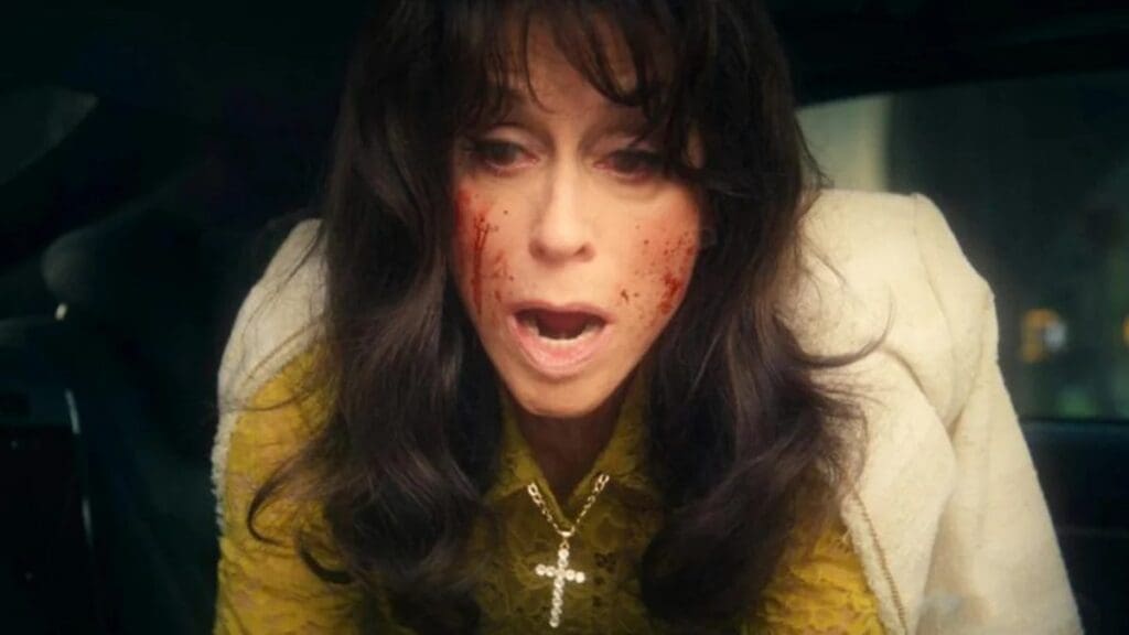 Woman with blood on her cheeks and an anguished, open-mouthed expression inside a car, wearing a yellow lace blouse and a cross necklace.