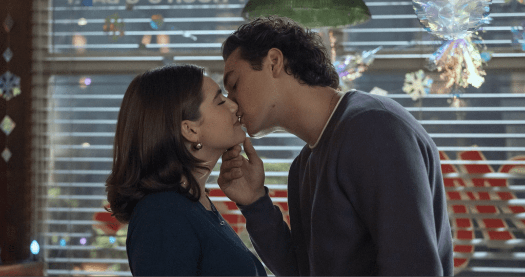A couple kissing indoors, the woman in a blue top and the man in a navy sweater, festive decorations in the background.