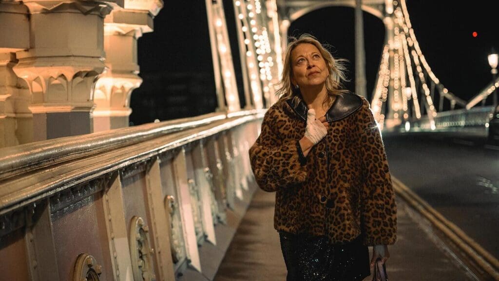 Woman in a leopard print coat stands on a brightly lit bridge at night, looking upward thoughtfully.