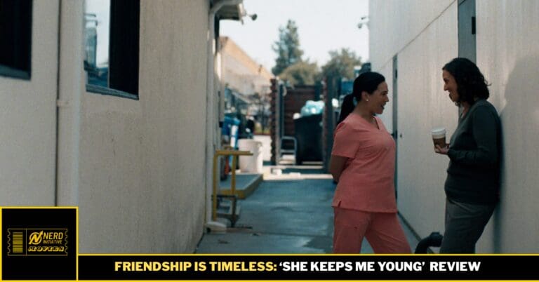 Two women chat and laugh in a narrow outdoor alley; one wears pink scrubs, the other holds a coffee cup and smiles at her.