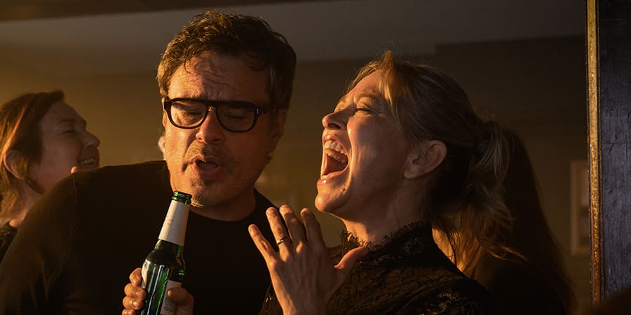 Man with glasses holds a beer while singing at a party, next to a woman who is also singing and laughing in warm indoor lighting in the background.