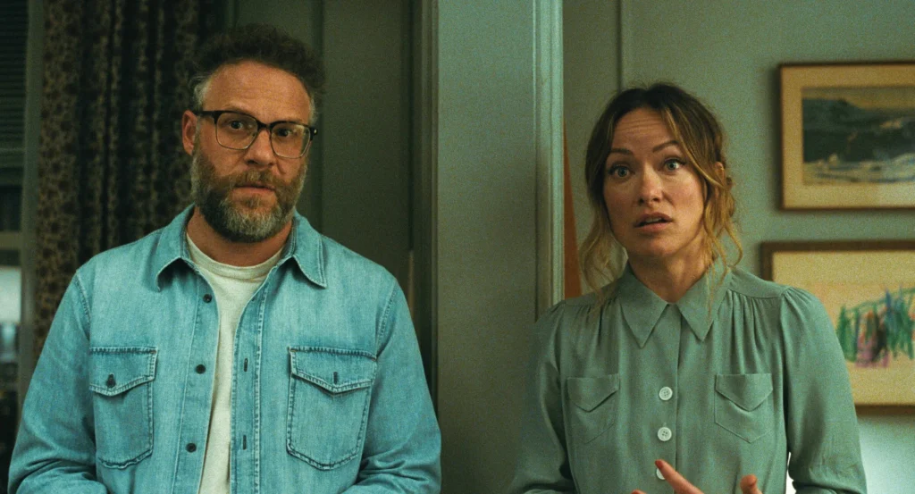 Man with glasses and beard in a denim shirt stands beside a surprised woman in a green blouse inside a room.