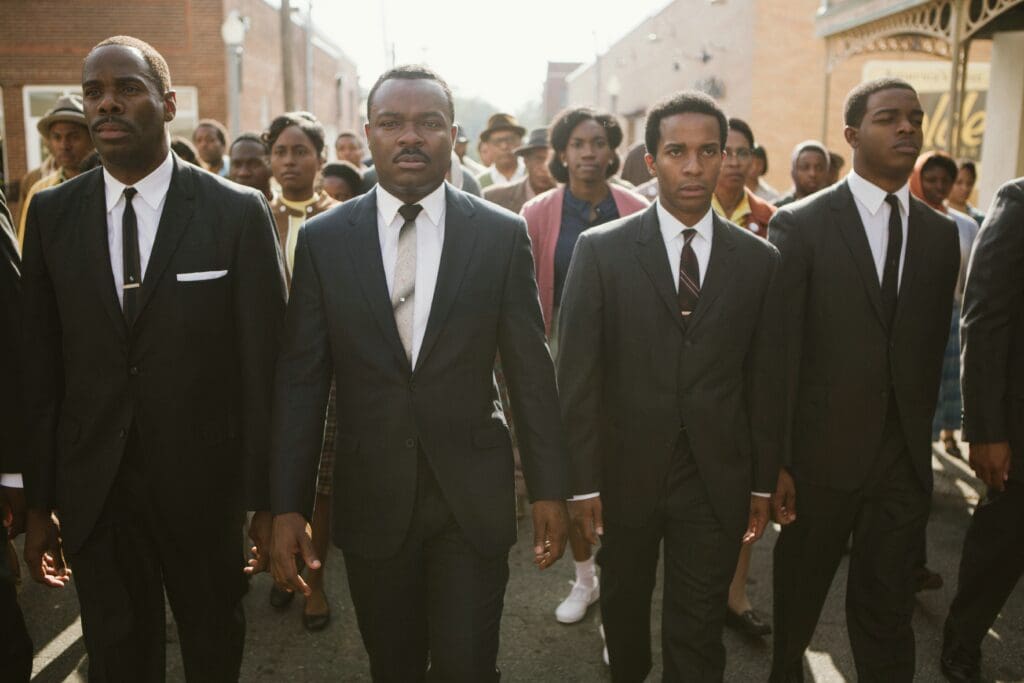 Group of men in black suits and ties walking down a city street, serious expressions in the foreground.