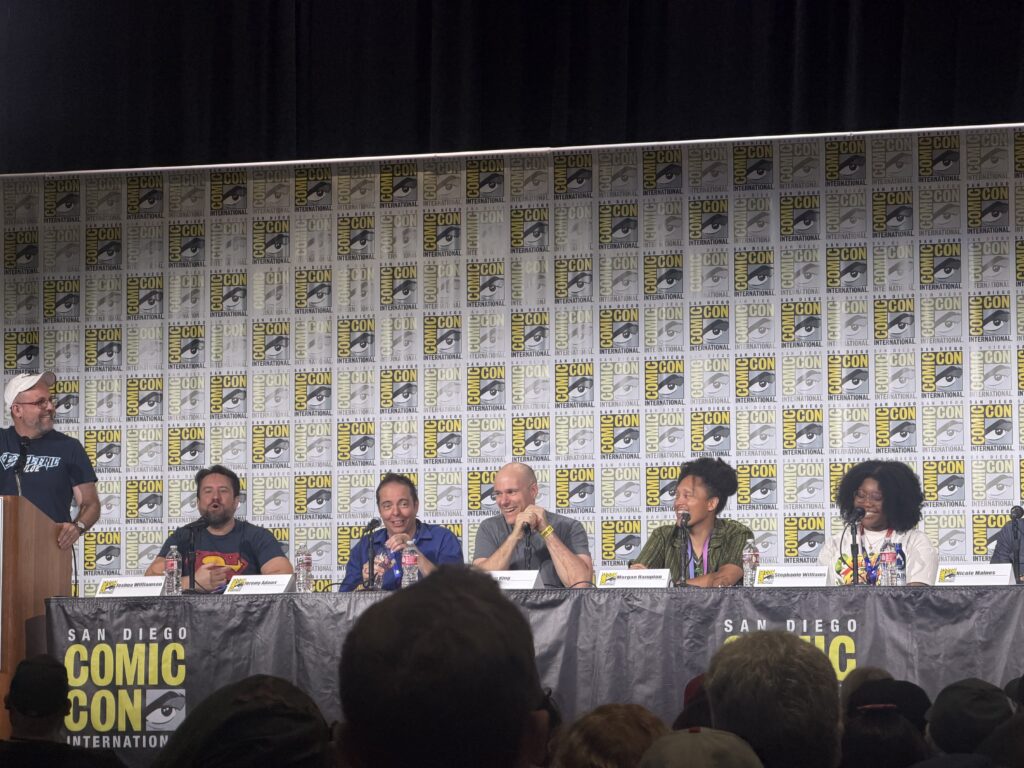 Panel of creators and speakers seated at a long table on stage, San Diego Comic-Con banners behind them.