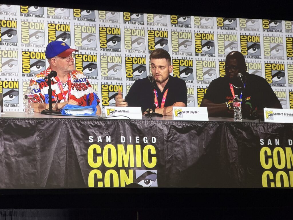 Three panelists sit at a table during a San Diego Comic-Con event, microphones and water bottles in front of them, backdrop filled with Comic-Con logos.