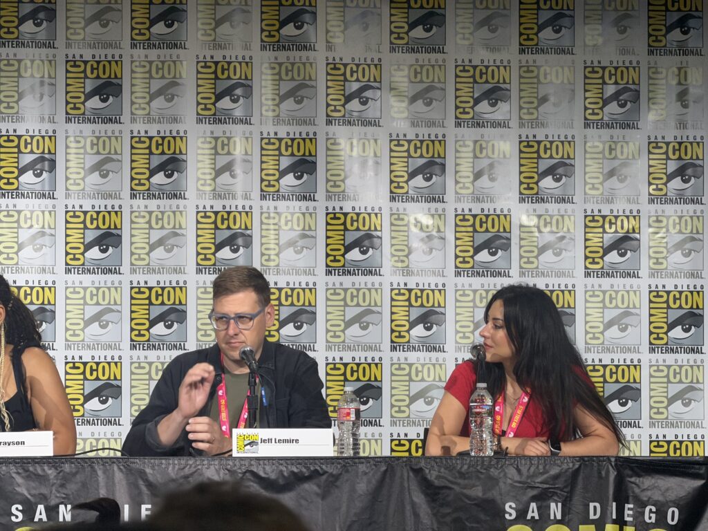 Panel at San Diego Comic-Con with three speakers seated behind a table; backdrop shows Comic-Con logos; center man with glasses speaks into a microphone; nameplate reads Jeff Lemire; woman on the right.