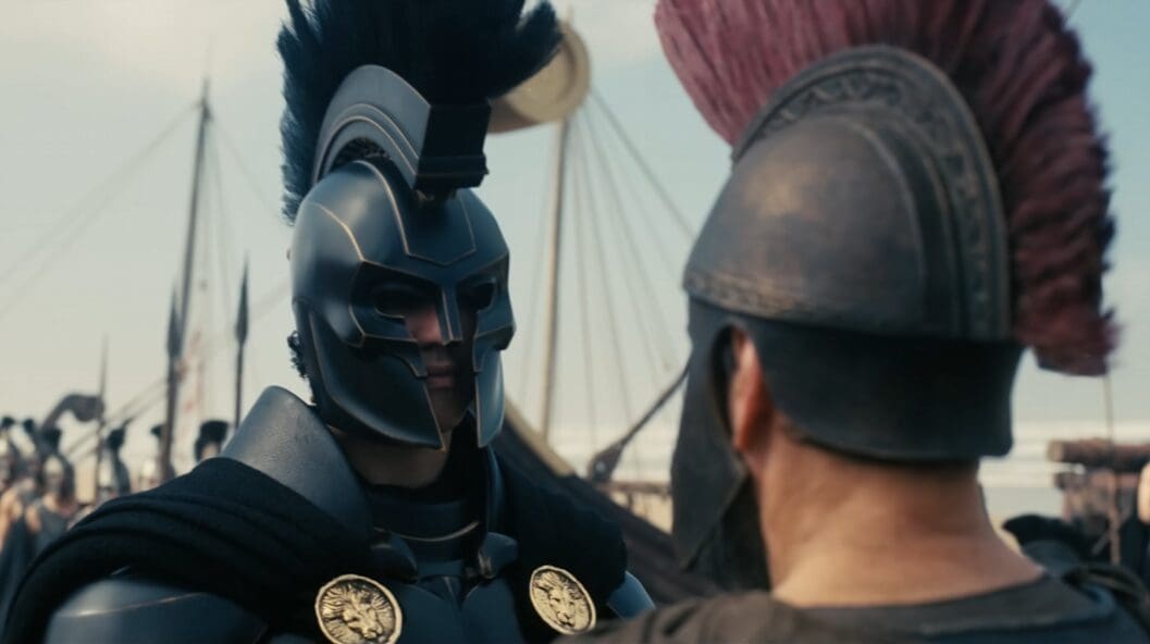 Two armored warriors face off; one wears a dark helmet with a crest and cloak, the other seen from behind with a plumed helmet at a reenactment scene