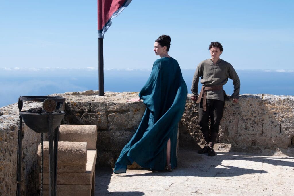 Two people in period clothing stand on a stone sea wall, with a red flag nearby and a brazier in the foreground against a blue sky and sea.