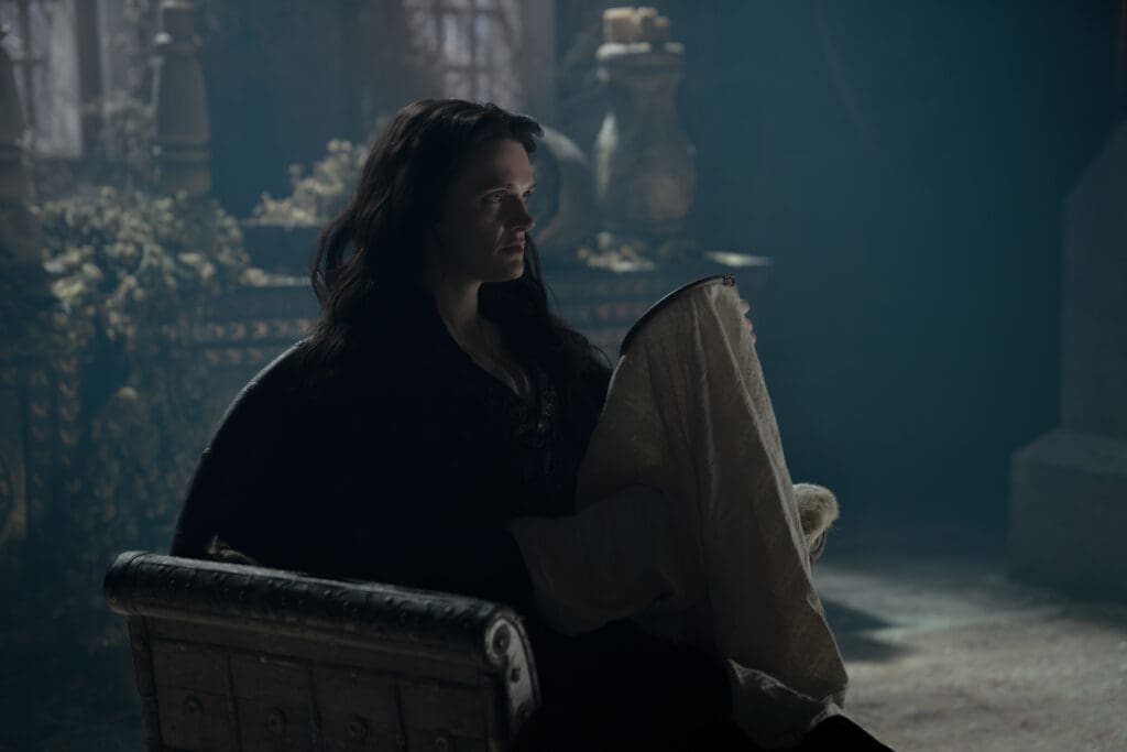 Woman sits inside a wooden chest, wrapped in a light blanket, in a dim blue-toned room.