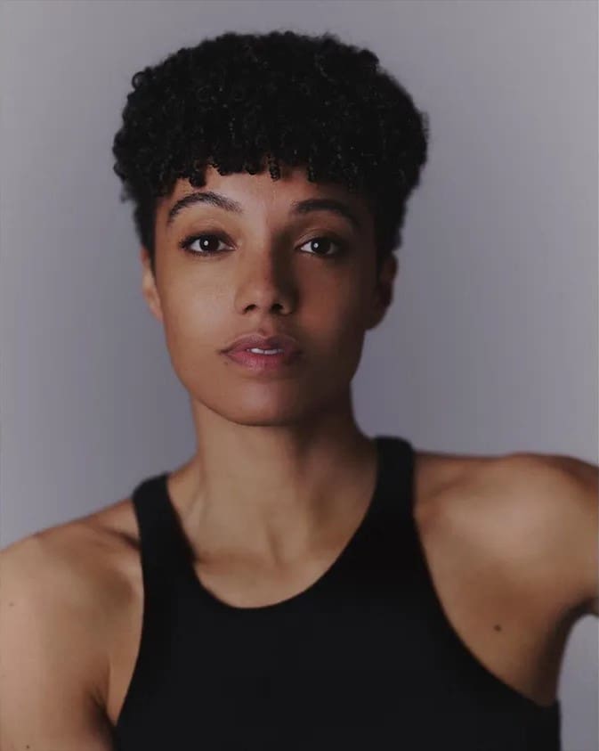 Portrait of a woman with short curly hair wearing a black tank top, looking at the camera against a neutral gray background.