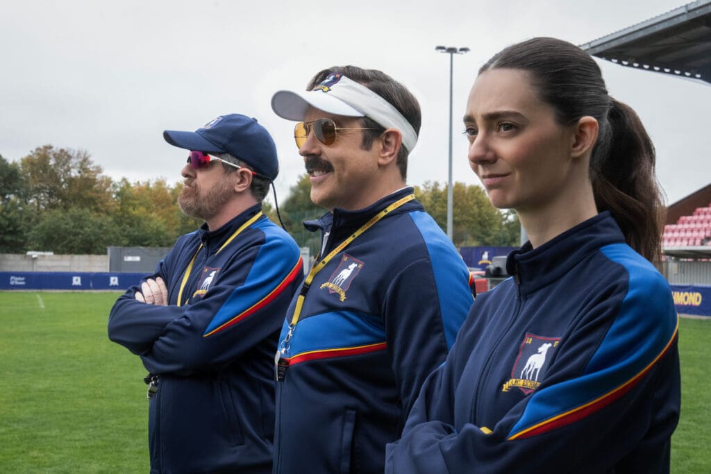 Three athletes in matching blue jackets with patches stand at a sports field, gazing to the left.
