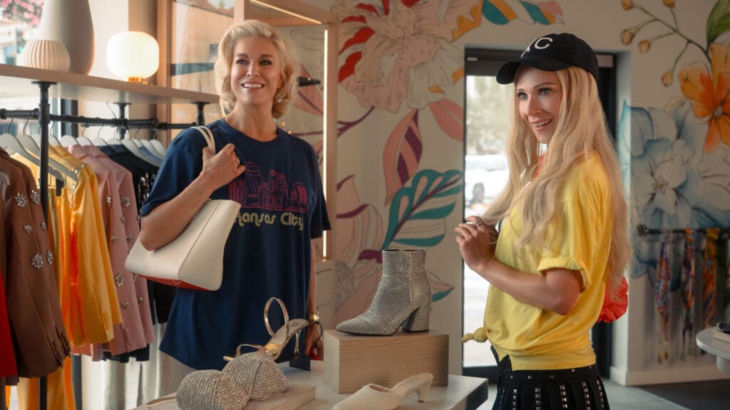 Two women in a bright fashion boutique smile at each other, with racks of colorful dresses behind them and glittery shoes on display.